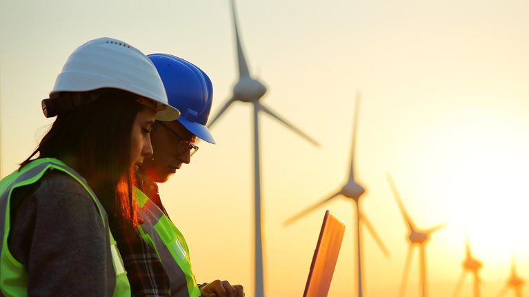 photo of two workers looking at laptop outside wind turbines with the backdrop of a beautiful sunset