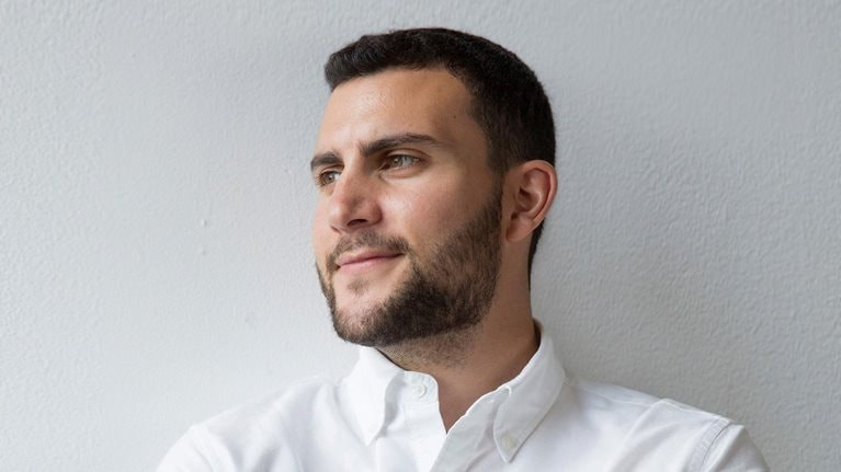 Portrait of Forage’s CEO Ofek Lavian. Lavian has a subtle smile and his face turned away from the camera as he looks off into the distance. He's wearing a white button down shirt and is leaning against a white textured wall.