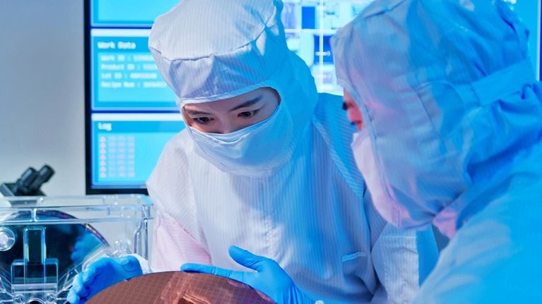Two Asian technicians in sterile coverall hold wafer with gloves and check it at semiconductor manufacturing plant