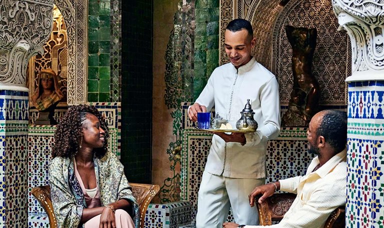 Smiling couple being served tea by concierge in courtyard of luxury hotel while on vacation