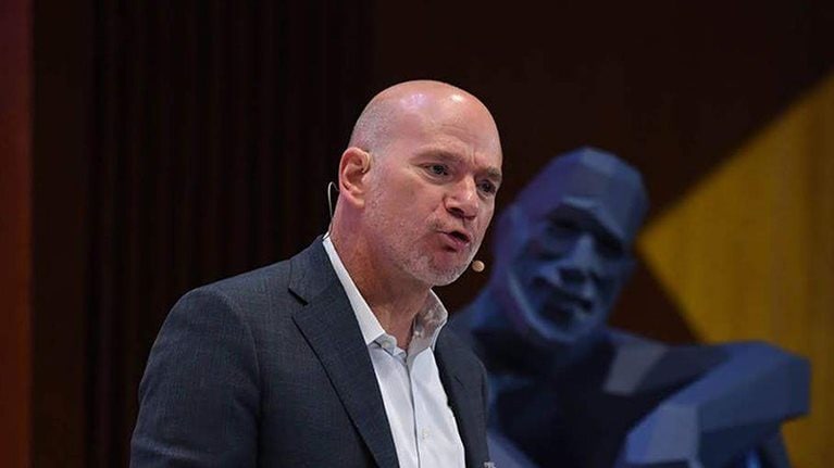 Andy McAfee speaking on stage with a smiling blue statue in a relaxed pose behind him.